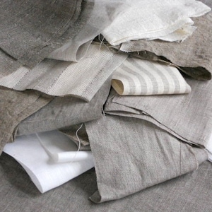 May include: A pile of different textured linen fabrics in shades of grey and white. The fabrics are folded and layered on top of each other.