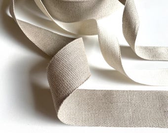 May include: A roll of beige woven fabric ribbon with a visible textured weave. The ribbon is partially unspooled, showing a flat, flexible length of material with finished edges. The light tan color is consistent throughout the entire length of the ribbon.