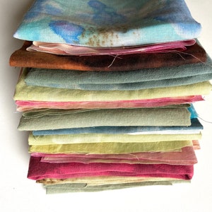 Op de afbeelding: Een stapel van 13 gevouwen vierkante stukken stof in verschillende tinten groen, blauw, geel, roze en bruin. De stof heeft een natuurlijke, handgekleurde uitstraling.
