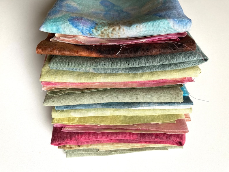 Op de afbeelding: Een stapel van 13 gevouwen vierkante stukken stof in verschillende tinten groen, blauw, geel, roze en bruin. De stof heeft een natuurlijke, handgekleurde uitstraling.