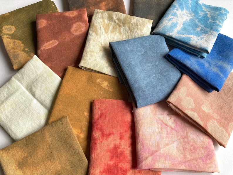 Op de afbeelding: Een verzameling van 14 vierkante stukken stof in verschillende tinten bruin, blauw, rood en roze. De stofvierkanten hebben een tie-dye-effect en zijn in een willekeurig patroon gerangschikt.