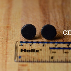 Puede incluir: Dos pendientes peque&ntilde;os, redondos y negros se muestran sobre una superficie de madera junto a una regla. Los pendientes tienen aproximadamente 1,3 cm de di&aacute;metro. La regla est&aacute; marcada en cent&iacute;metros.