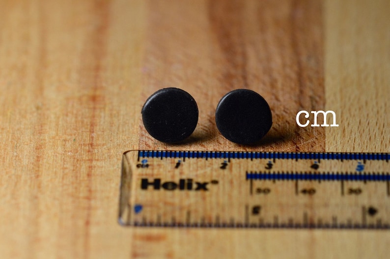Puede incluir: Dos pendientes peque&ntilde;os, redondos y negros se muestran sobre una superficie de madera junto a una regla. Los pendientes tienen aproximadamente 1,3 cm de di&aacute;metro. La regla est&aacute; marcada en cent&iacute;metros.