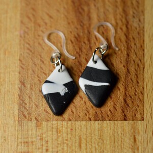 May include: A pair of diamond-shaped earrings with a black and white pattern. The earrings have a silver-colored metal loop and a clear plastic ear wire. The black and white design is abstract, with the white sections appearing to be painted on.