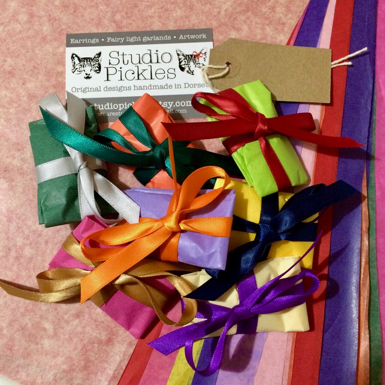 Puede incluir: Una colecci&oacute;n de peque&ntilde;os regalos envueltos en papel de colores y atados con cintas de raso. Los regalos son de varios colores, incluyendo verde, naranja, morado y amarillo. Se ve una tarjeta de visita con el texto "Studio Pickles".