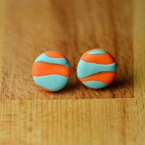 May include: A pair of round earrings with an orange and light blue striped design. The earrings are set against a wooden background.