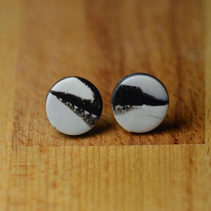 May include: A pair of round stud earrings. Each earring features a white background with a black, abstract design. The earrings are set against a wooden surface.