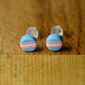 May include: Two round stud earrings featuring a light blue base with horizontal white and pink stripes. The earrings are displayed on a wooden surface, with clear earring backs visible behind them.