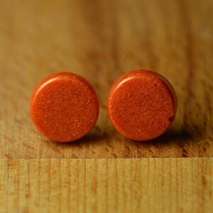 May include: A pair of circular stud earrings featuring a vibrant orange hue with a subtle, fine-grained glittery texture throughout the surface. The earrings have a smooth, rounded edge and a flat face, presenting a consistent, uniform appearance.
