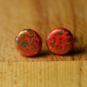May include: Two round, red earrings with gold flecks. The earrings are set against a wooden background. The earrings are a vibrant red color with irregular gold leaf patterns, creating a unique and eye-catching design.