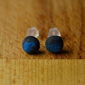 May include: A pair of round stud earrings featuring a dark charcoal grey base with a textured, speckled blue finish on the front surface. Each earring includes a clear, translucent plastic backing post attached to the rear of the circular stud.