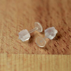 May include: Two clear, translucent plastic earring backs with a flat circular disc base and a small cylindrical post. These small jewelry components are designed to secure stud earrings in place, featuring a soft, flexible material construction.