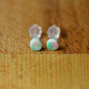 May include: Handmade stud earrings with a marbled effect. One earring has a mix of white, light blue, and green, the other pink and green. Clear earring backs are also visible, set against a wooden surface.