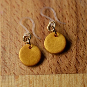 May include: A pair of gold disc earrings with clear plastic ear wires. The earrings feature a simple design, with a small, round gold disc suspended from a gold-coloured hook.