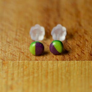 May include: A pair of stud earrings featuring small, round, flat-faced circular tops. Each earring is divided into two distinct halves, one coloured in a vibrant lime green and the other in a deep purple, complete with clear plastic earring backs.