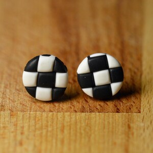 May include: A pair of round stud earrings featuring a black and white checkered pattern. Each earring has a circular shape with a grid of alternating black and white squares, creating a distinct geometric design on the front surface.