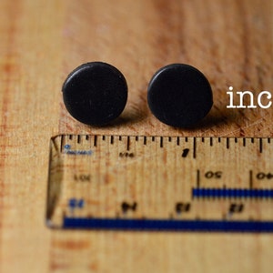 Puede incluir: Dos pendientes peque&ntilde;os, redondos y negros se muestran sobre una superficie de madera junto a una regla. La palabra "inches" est&aacute; escrita en texto blanco. Los pendientes tienen aproximadamente 1,3 cm de di&aacute;metro.