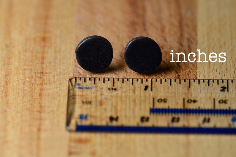 Puede incluir: Dos pendientes peque&ntilde;os, redondos y negros se muestran sobre una superficie de madera junto a una regla. La palabra "inches" est&aacute; escrita en texto blanco. Los pendientes tienen aproximadamente 1,3 cm de di&aacute;metro.