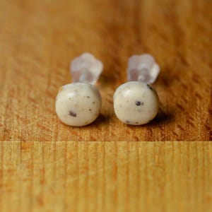 Peut inclure: Une paire de boucles d'oreilles à clous rondes présentant une surface en céramique blanc cassé lisse avec de petites taches gris foncé et brunes dispersées. Chaque boucle possède un fermoir en plastique translucide fixé à l'arrière du clou.