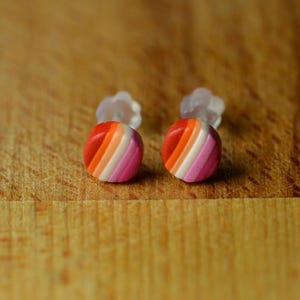 Peut inclure: Deux boucles d'oreilles rondes avec un motif rayé rouge, orange, blanc, rose et violet. Les boucles d'oreilles sont posées sur une surface en bois, avec des fermoirs transparents visibles.