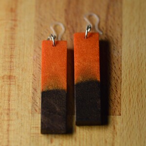 May include: A pair of rectangular earrings with a gradient design. The top half is orange, transitioning to black at the bottom. The earrings have silver-colored hooks and clear ear wires. The earrings are displayed on a wooden surface.