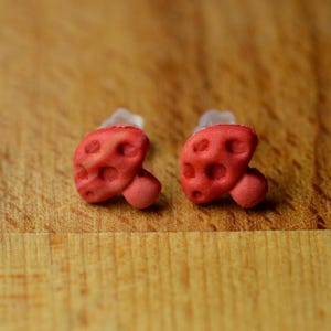 May include: A pair of stud earrings shaped like small red mushrooms. Each earring features a textured, rounded cap with indented spots and a short, cylindrical stem. The earrings are attached to clear plastic earring backs for secure wear.