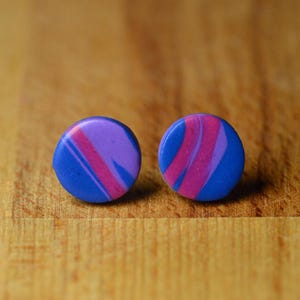 May include: A pair of round stud earrings with a marbled design. The earrings feature a blue base with streaks of purple and pink. The earrings are set against a wooden surface.
