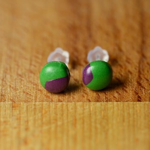 May include: A pair of stud earrings featuring small, round, green spheres with a distinct purple patch on each. The earrings are equipped with clear, translucent plastic earring backs, providing a simple and functional design for these circular accessories.
