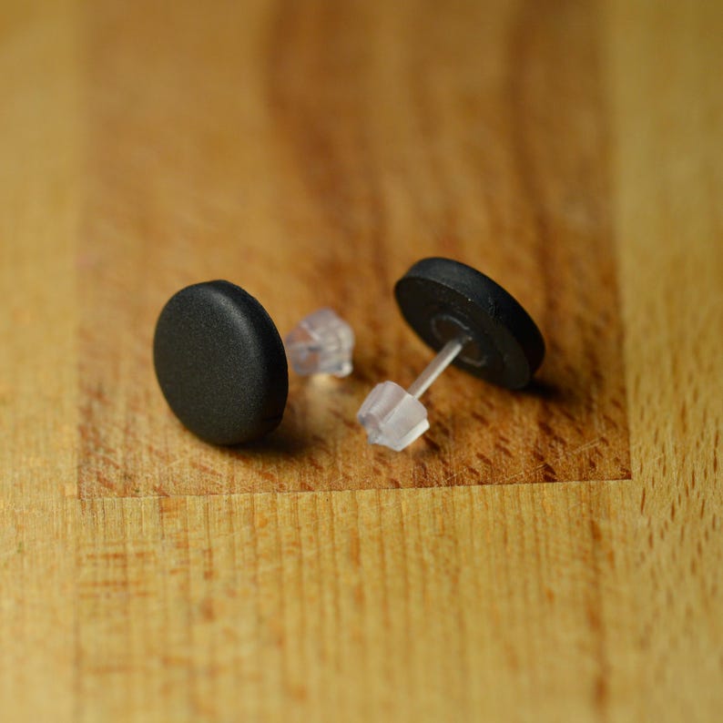 May include: A pair of circular stud earrings featuring a matte black finish on the front face. Each earring includes a clear, translucent plastic post and a matching clear plastic butterfly-style earring back for secure attachment to the ear.
