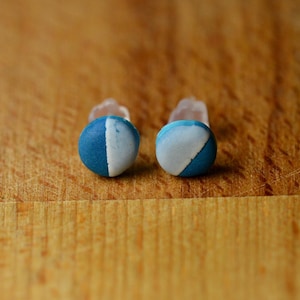 May include: A pair of round stud earrings. Each earring is a small sphere, split diagonally with a white and blue color scheme. The earrings are set on clear plastic posts, resting on a wooden surface.