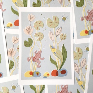 May include: A light blue background with a whimsical illustration of two pink frogs jumping in a pond with lily pads, snails, and bugs. The pond is surrounded by green lily pads and flowers.