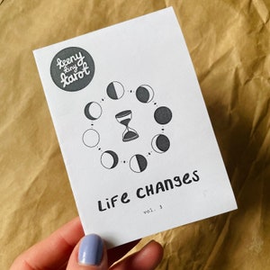 May include: A white card with black text and illustrations. The text reads "teeny tiny tarot" and "Life Changes vol. 3". The card features a circle of moons and a black hourglass.