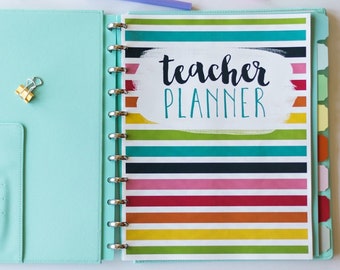May include: A teal teacher planner with colorful stripes and the text "teacher planner" in white lettering. The planner has a ring binding and a tabbed divider.
