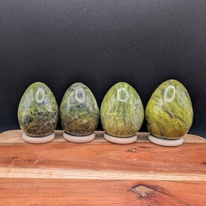 May include: Four green stone eggs with natural veining patterns, each resting on a small, round wooden stand. The eggs vary in shades of green and yellow, creating a natural, earthy aesthetic. The background is a solid black.