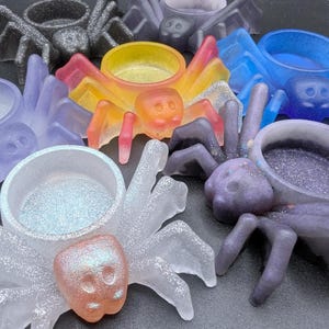 May include: A collection of colorful spider-shaped resin containers. The spiders are in various colors, including orange, yellow, blue, purple, and clear, with glitter accents. Each spider has a circular bowl on its back.
