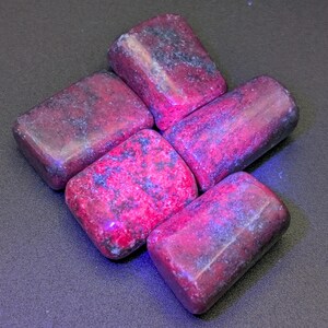 May include: Six tumbled stones of red ruby in various shapes and sizes. The stones are arranged in a cluster on a dark gray surface.