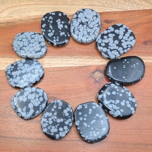 May include: A collection of oval-shaped snowflake obsidian stones. The stones are black with white or gray snowflake-like patterns. The stones are arranged in a circular pattern on a wooden surface.