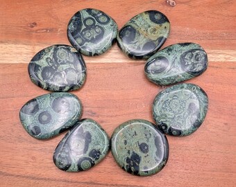 May include: Eight polished, oval-shaped stones arranged in a circle. The stones are primarily green with dark, circular patterns. The stones are on a wooden surface.