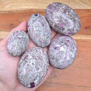 May include: Five oval-shaped stones with a speckled pattern of gray and purple hues. The stones vary in size and are held in a hand, showcasing their natural textures and colors. The stones have a polished appearance.