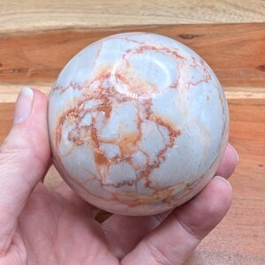 May include: A marble sphere with a light blue base color and intricate reddish-brown veining. The polished stone ball is held in a hand, showcasing its smooth surface and natural patterns. This decorative sphere is a unique home decor item.
