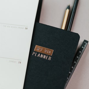 May include: A black planner with the words "MY BIG PLANNER" in copper lettering. A white notebook is open next to it, with the date "2021" visible. Two pencils and a ruler are tucked inside the planner. Rose gold paper clips are in the background.