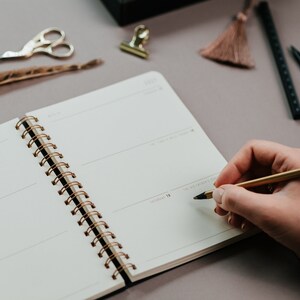 May include: A person writing in a spiral-bound notebook with a gold pen. The notebook is open to a blank page with lines for writing. Other stationery items include gold scissors, a feather, and a tassel.