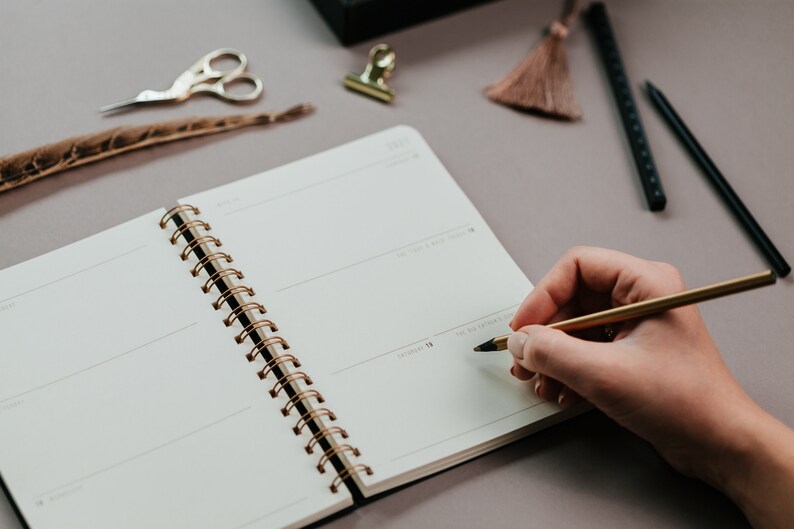 May include: A person writing in a spiral-bound notebook with a gold pen. The notebook is open to a blank page with lines for writing. Other stationery items include gold scissors, a feather, and a tassel.