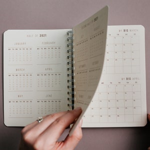 May include: Open spiral-bound planner with a neutral color scheme. The planner is open to display the calendar pages for the second half of 2021. The pages are being turned to reveal the months of July, August, September, October, November, and December.