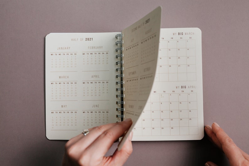 May include: Open spiral-bound planner with a neutral color scheme. The planner is open to display the calendar pages for the second half of 2021. The pages are being turned to reveal the months of July, August, September, October, November, and December.