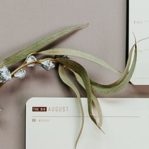 May include: A close-up of a calendar page with the text "THE BIG AUGUST" and the days of the week. A sprig of eucalyptus with green leaves and white accents is placed on the calendar. The background is a muted purple.