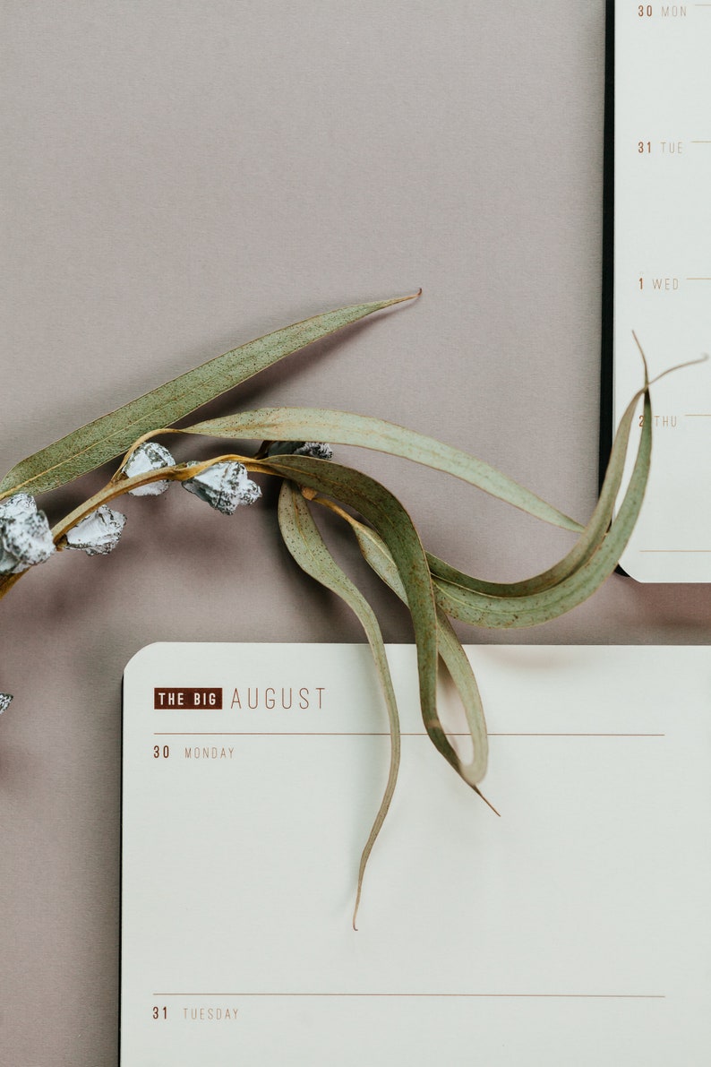 May include: A close-up of a calendar page with the text "THE BIG AUGUST" and the days of the week. A sprig of eucalyptus with green leaves and white accents is placed on the calendar. The background is a muted purple.