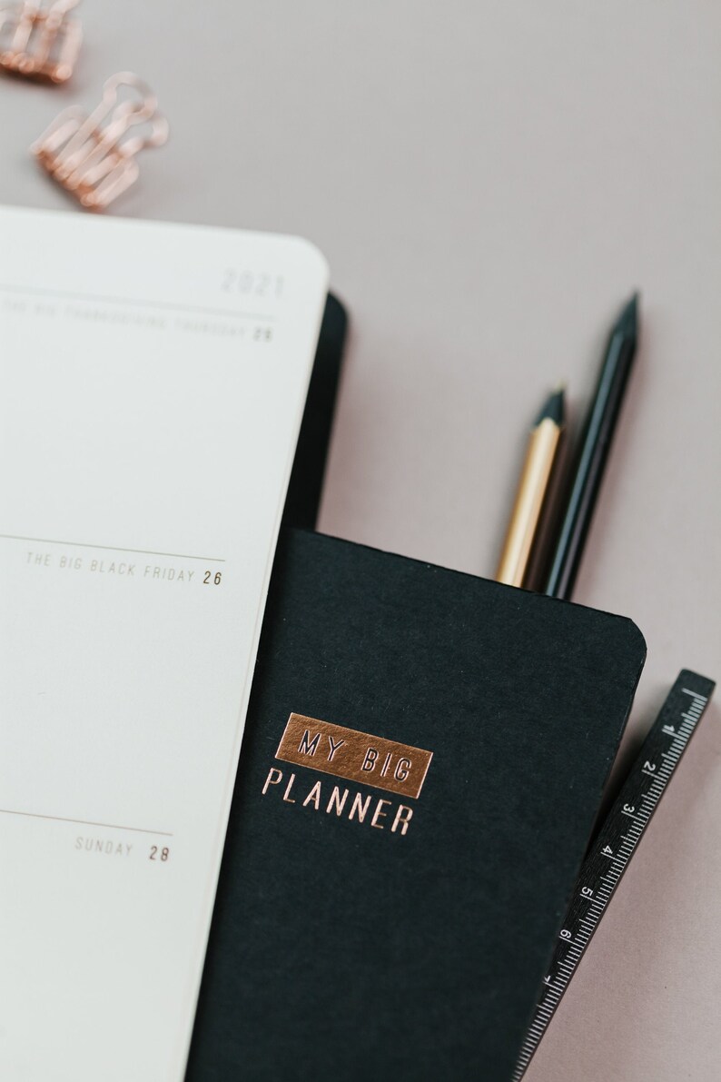 May include: A black planner with the words "MY BIG PLANNER" in copper lettering. A white notebook is open next to it, with the date "2021" visible. Two pencils and a ruler are tucked inside the planner. Rose gold paper clips are in the background.