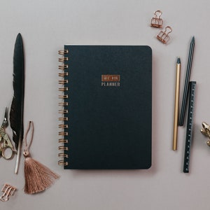May include: A flat lay featuring a dark teal planner with gold spiral binding and the text "MY BIG PLANNER". Accompanying the planner are gold and black pens, a ruler, paper clips, a decorative alligator, a feather, scissors, and a tassel, all arranged on a neutral surface.