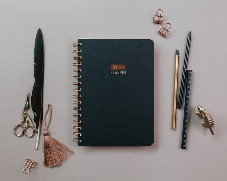 May include: A flat lay featuring a dark teal planner with gold spiral binding and the text "MY BIG PLANNER". Accompanying the planner are gold and black pens, a ruler, paper clips, a decorative alligator, a feather, scissors, and a tassel, all arranged on a neutral surface.